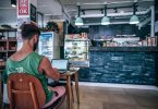 ein cafe mit einer schwarzen Bar, links sitz auf einem Holzstuhl ein junger Mann mit grünem t-Shirt und arbeitet an einem kleinem Laptop