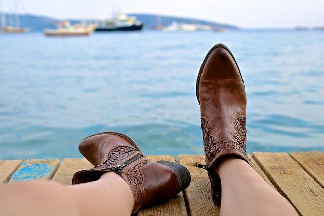 die beine einer Person die am Steg sitzt und aufs Meer schaut mit braunen Cowboystiefeln an den Füßen