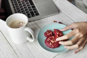 Ausschnitt eines weissen Holztisches auf dem ein Laptop, eine halb volle weisse Kaffeetasse und ein kleiner grüner runder Teller mit red velvet Keksen stehen, eine feminine Hand nimmt sich gerade einen Keks