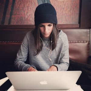 eine junge Frau mit schwarzer Mütze und grauem Pullover sitzt auf einer Lederbank und arbeitet an einem grossen Apple-Laptop
