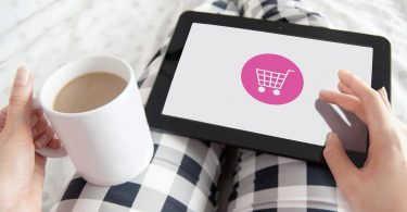 eine Person mit schwarz-weiß karierter Hose die mit im Bett liegt in der linken Hand eine Tasse Milchkaffee hält und mit der rechten Hand auf einem Tablet online shoppt