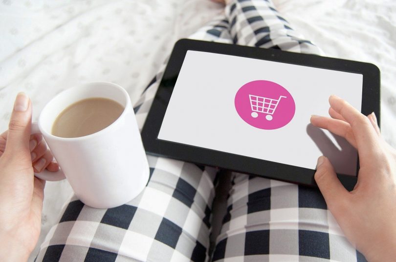 eine Person mit schwarz-weiß karierter Hose die mit im Bett liegt in der linken Hand eine Tasse Milchkaffee hält und mit der rechten Hand auf einem Tablet online shoppt