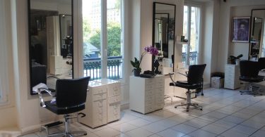 ein Friseursalon in weiß mit schwarzen Sesseln die vor spiegeln sehen mit einer kleinen weißen Kommode daneben und Fenstern dazwischen