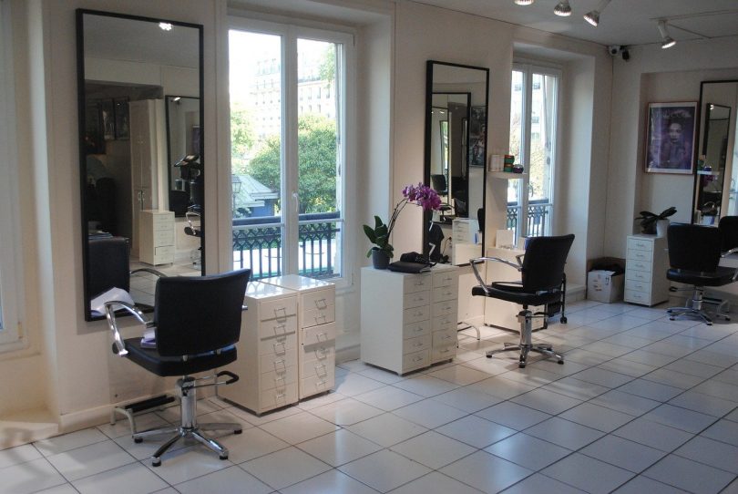 ein Friseursalon in weiß mit schwarzen Sesseln die vor spiegeln sehen mit einer kleinen weißen Kommode daneben und Fenstern dazwischen