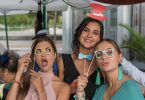 vier junge Frauen die verschieden assecoirs wie Brillen, Hütte oder Fliegen an einem Stock halten und für das Foto lächeln