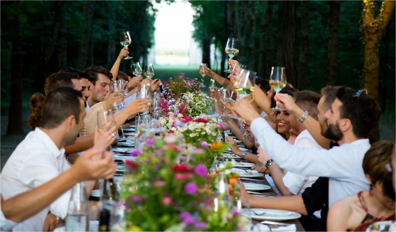 eine langer gedeckter Tisch unter freiem Himmel an dem viele Leute sitzen die gerade ihr Glas zum anstoßen hoch heben