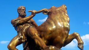 eine bronzene Statue von Herkules wie er auf einem Stier reitet, dahinter blauer Himmel mit vereinzelten Wolken