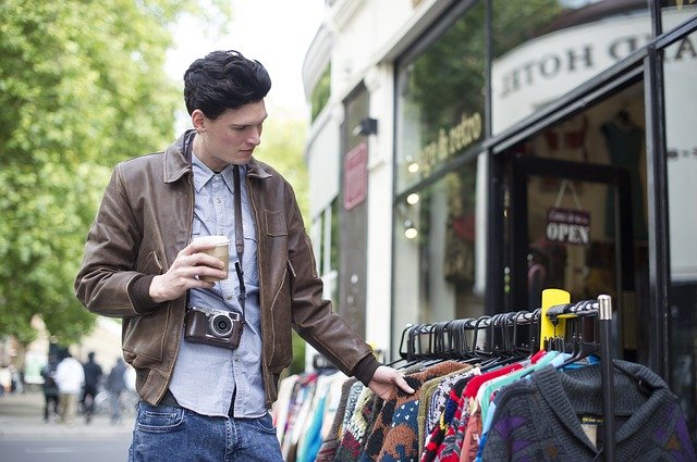 ein junger Mann mit einem coffee to go in der Hand der durch den Kleiderständer vor der Tür eines Second Hand Ladens stöbert