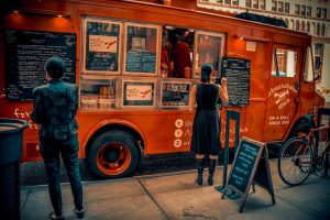 ein rötlicher Foodtuck mit vielen Tafeln auf der mit weisser Kreide alle Gerichte aufgelistet sind, eine junge Frau im schwarzen Kleid gibt gerade eine Bestellung auf und eine andere schaut sich das Menu an