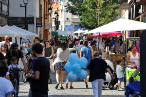 Strasse mit Menschen und weissen Sonnenschirmen in der Mitter eine Frau mit Kind das sechs hellblaue Luftballons hinterher zieht