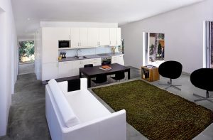 H Arquitectes, ein Loft, im Vordergrund ein weisses Sofa und zwei schwarze moderne Sessel gegenüber, in der Mitter ein brauner Esstisch mit vier braunen Stühlen und ganz hinten eine weisse Küchenzeile