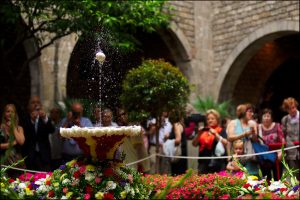 eine Menschenmenge die um einen Springbrunnen im Vorhof einer Kirche steht und ein weißes Ei auf seinem Wasserstrahl tanzen lässt, drum herum ist alles mit Blumen geschmückt