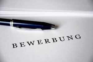 eine weiße Mappe auf der in schwarzen Großbuchstaben Bewerbung steht und darüber liegt ein blauer Kugelschreiber
