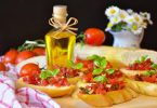 ein Tisch mit sechs Tomatenbroten, eine kleine Flasche mit Olivenöl und im Hintergrund ein angeschnittenes Baguette, Tomaten und eine Vase mit Gänseblümchen