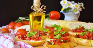 ein Tisch mit sechs Tomatenbroten, eine kleine Flasche mit Olivenöl und im Hintergrund ein angeschnittenes Baguette, Tomaten und eine Vase mit Gänseblümchen