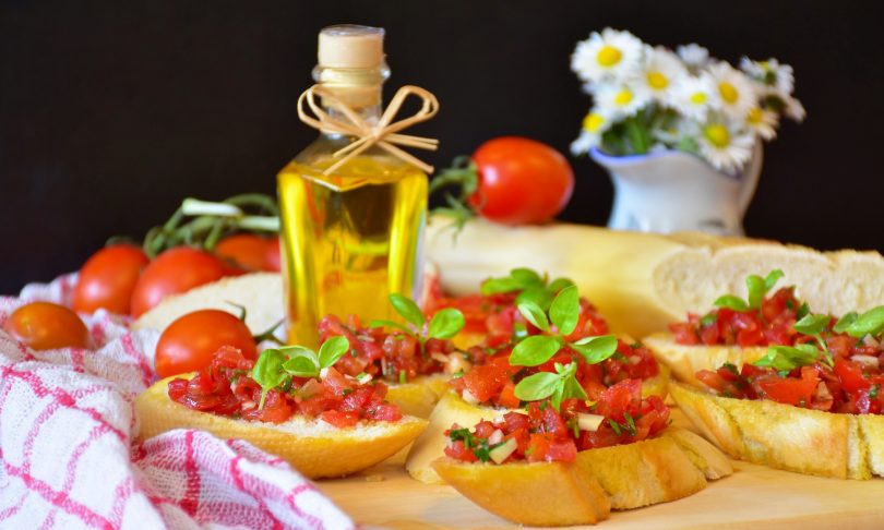 ein Tisch mit sechs Tomatenbroten, eine kleine Flasche mit Olivenöl und im Hintergrund ein angeschnittenes Baguette, Tomaten und eine Vase mit Gänseblümchen