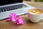 Bogambillablüten zwischen einem Milchkaffe mit Herzmuster im Schaum und man sieht einen Teil eines Laptops auf einem Holztisch