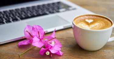 Bogambillablüten zwischen einem Milchkaffe mit Herzmuster im Schaum und man sieht einen Teil eines Laptops auf einem Holztisch