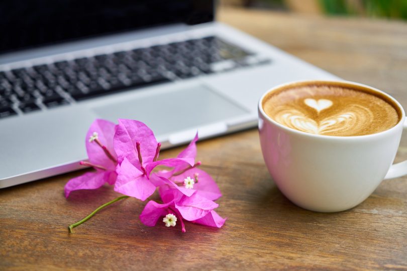 Bogambillablüten zwischen einem Milchkaffe mit Herzmuster im Schaum und man sieht einen Teil eines Laptops auf einem Holztisch