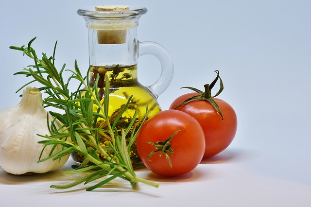 ein kleines Fläschchen Olivenöl, davor eine Knoblauchzehe, ein kleiner Rosmarizweig und daneben zwei kleine Tomaten