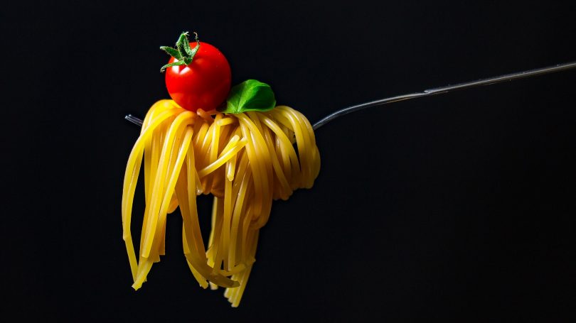 eine Gabel mit aufgerollten Spaghetti einer Kirschtomate darauf und ein Basilikumblatt, schwarzer Hintergrund