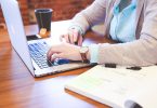 eine Frau mit hellblauer bluse und graue Strickjacke die an einem Holztisch sitzt und an einem laptop schreibt, rechts von ihr eine Tasse Tee und links ein offenes buch