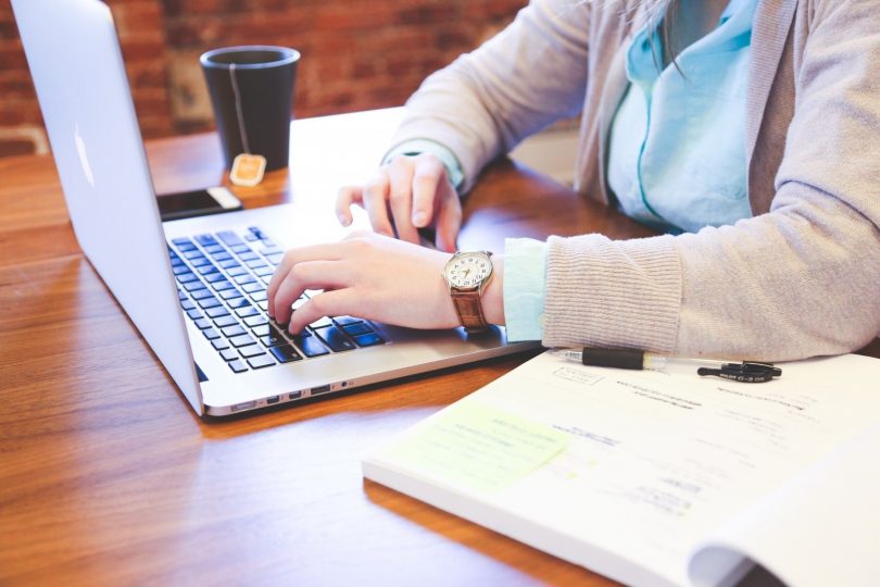 eine Frau mit hellblauer bluse und graue Strickjacke die an einem Holztisch sitzt und an einem laptop schreibt, rechts von ihr eine Tasse Tee und links ein offenes buch