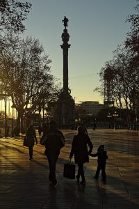 im Hintergrund die Kolumbusstatue bei Sonnenuntergang und im Vordergrund eine Familie mit einem kleinen Kind, alle warm eingepackt und die Frau hält eine Einkaufstüte in der linken Hand