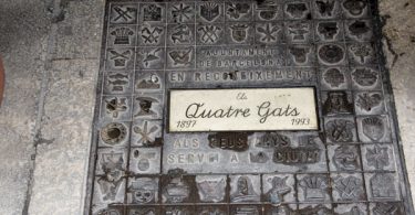 eine metallplatte auf dem Boden die mit verschiedenen Zeichen dekoriert ists und in der Mitte steht in Schreibschrift Qutatre Gats