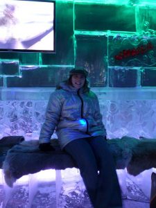 Eine Frau mit dickem silbernen Anorak und Mütze die auf einer der Eisbänke mit Fell darauf sitzt, dahinter ist ein Fernseher zu sehen und das eis spiegelt grünes und lilanes Licht wieder