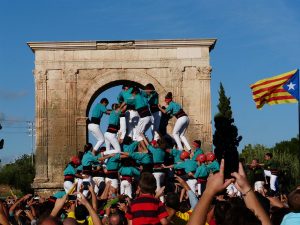 der Aufbau einer Menschpyramide der Catellers mit weissen Hosen und Türkisen T-Shirts, drum herum viele Leute die sie anfeuern und rechts im Bild eine katalanische Unabhängigkeitsflagge