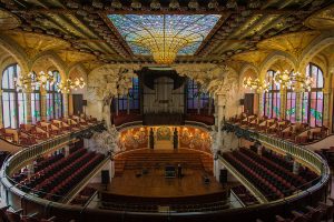 Der Palau de la Música Catalana von Innen, die Sitzreihen sind in ovaler Form um und über die Bühne verteilt, an den Seiten gewölbte Fenster und an der Decke ein buntes Glasmosaik mit einer imposanten Beleuchtung