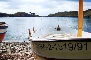ein Fischerboot am Strand einer Bucht die fast komplett von Bergen umzingelt ist, auf dem beigen Boot steht 7aBA-2-1519/91