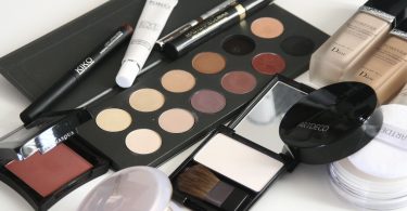 verschieden Kosmetika die auf einem weissen Untergrund quer durcheinander liegen, zum Beispiel eine Palette mit elf Brauntönen für Lidschatten, Pinsel, Bräunungspuder und Flüssig Make-Up