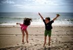 Ein junge mit grüner Badehose und blauem T-Shirt und ein Mädchen mit rosanem Badeanzug die am Strand springen und lachen