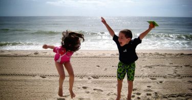 Ein junge mit grüner Badehose und blauem T-Shirt und ein Mädchen mit rosanem Badeanzug die am Strand springen und lachen
