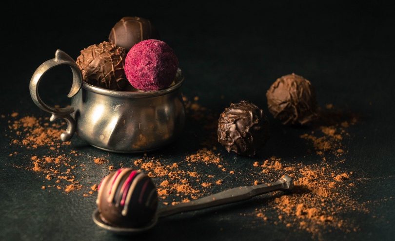 im Vordergrund ein Metalllöffel mit einer runden Schokoladenpraline rechts dahinter zwei weitere Pralinen, eine mit dunkler und eine mit Milchschokolade und links dahinter ein Metallbecher mit drei Pralinen eine davon ist rosa