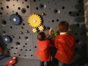 eine graue Wand mit gelben und blauen Zahnrädern in verschiedenen Größen an dem zwei kleine rotgekleidete Kinder spielen