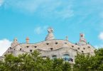 Aussicht von unten nach oben auf das Casa Mila mit grünen Bäumen davor und blauem Himmel darüber