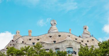 Aussicht von unten nach oben auf das Casa Mila mit grünen Bäumen davor und blauem Himmel darüber