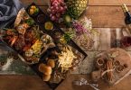 ein Holztisch auf dem ein Tablett mit Brot, Pommes, verschiedenen Sossen, Fleisch und Gemüse seht, recht oben ein Korb mit Obst und rechts unten zwei aufeinanderliegende Gabeln und ein kleines Holzbrett mit Zimtstangen und Orangenscheiben