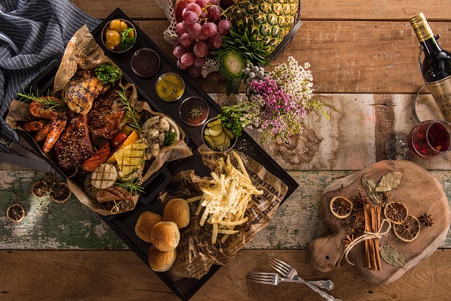 ein Holztisch auf dem ein Tablett mit Brot, Pommes, verschiedenen Sossen, Fleisch und Gemüse seht, recht oben ein Korb mit Obst und rechts unten zwei aufeinanderliegende Gabeln und ein kleines Holzbrett mit Zimtstangen und Orangenscheiben