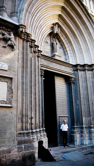 der imponente Eingang der Kathedrale Barcelonas, nur ein Türflügel ist geöffnet und vor dem Geschlossenen steht ein Mann mit schwarzer Hose und weissem Hemd