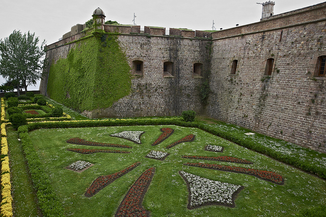 Teil der Mauern des Schlosses mit einem Vorgarten auf dem ein abstraktes Symbol durch Bepflanzung zu sehen ist
