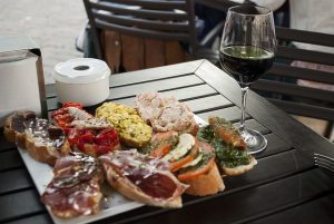 Terrassenholztisch auf dem ein weisser Aschenbecher, ein halb volles Weinglas und ein weisser viereckiger Teller mit verschiedenen Tapas auf Baguette steht