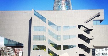 im Vordergrund ist das Museum Disney mit einer großen Terrasse davor mit Stühlen und Leuten die die Sonne genießen und hinter dem Gebäude sieht man die Spitze der Torre Agbar