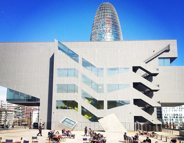 im Vordergrund ist das Museum Disney mit einer großen Terrasse davor mit Stühlen und Leuten die die Sonne genießen und hinter dem Gebäude sieht man die Spitze der Torre Agbar