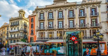 Aufzug einer Metrohaltestelle auf einem Platz in Barcelona, dahinter fährt ein torquier Bus vorbei und an der Seite ist eine Terrasse eines Cafés mit beigen Sonnenschirmen