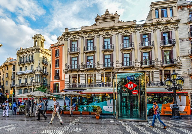 Aufzug einer Metrohaltestelle auf einem Platz in Barcelona, dahinter fährt ein torquier Bus vorbei und an der Seite ist eine Terrasse eines Cafés mit beigen Sonnenschirmen