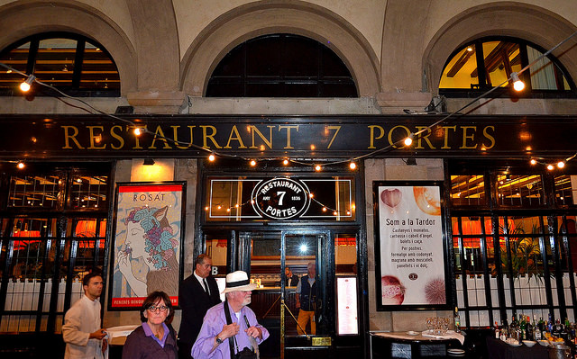 die Fasade des Restaurants 7 Portas mit schwarzen Fensterrahmen und goldener Schrift, einer Lichterkette davor und ein Kellner mit Krawatte an der Tür